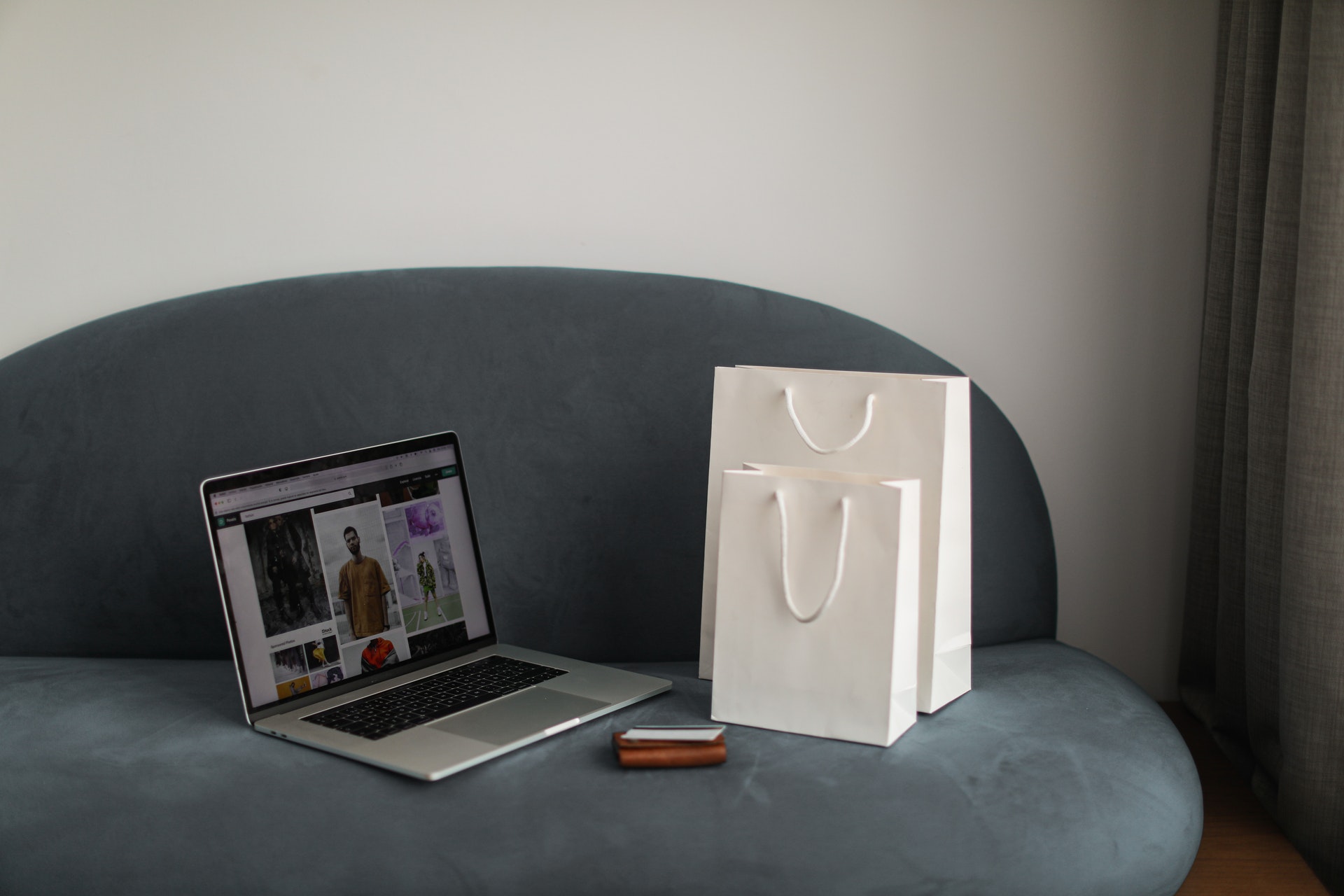 Grey couch with an open laptop, a phone in a brown pouch and two 
                        white shopping of different sizes on top of it.